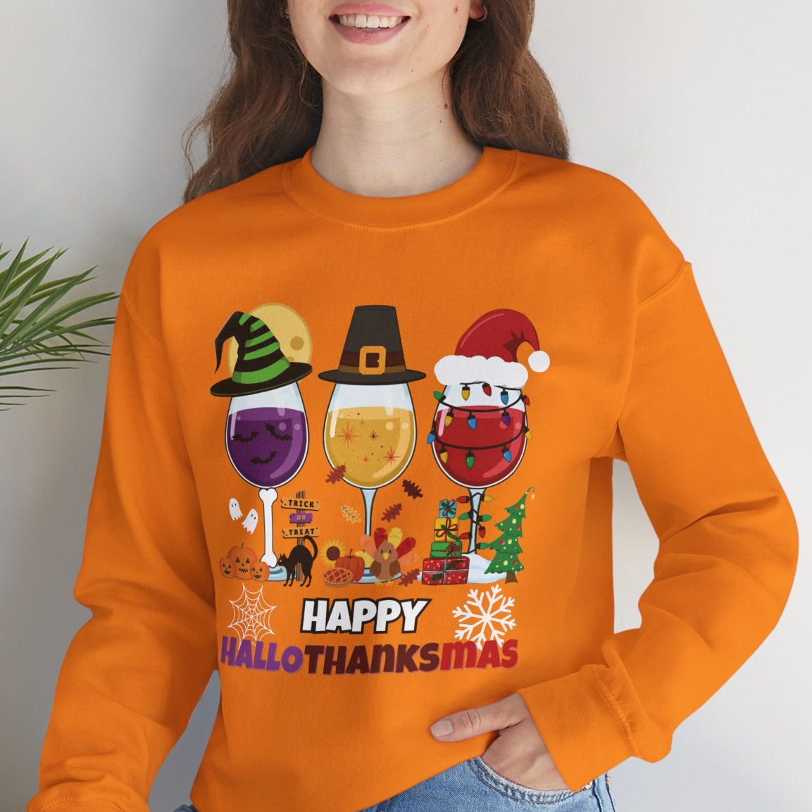Person wearing an orange sweatshirt with 'Happy HalloThanksMas' design, sitting on a white surface with a plant in the background.