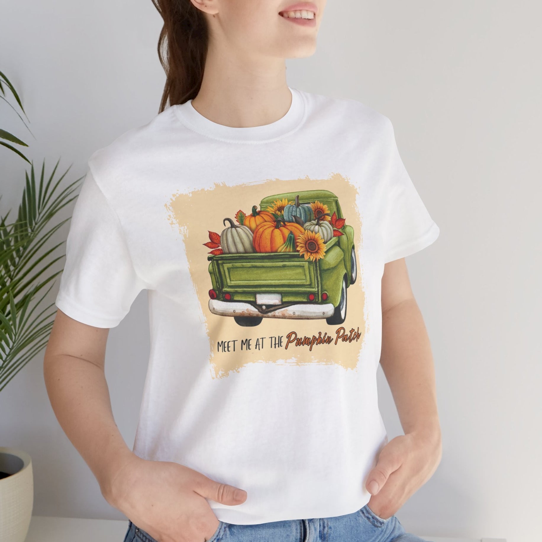 Person wearing a white t-shirt with a graphic of a green truck filled with pumpkins and fall decorations, text 'Meet me at the Pumpkin Patch'.