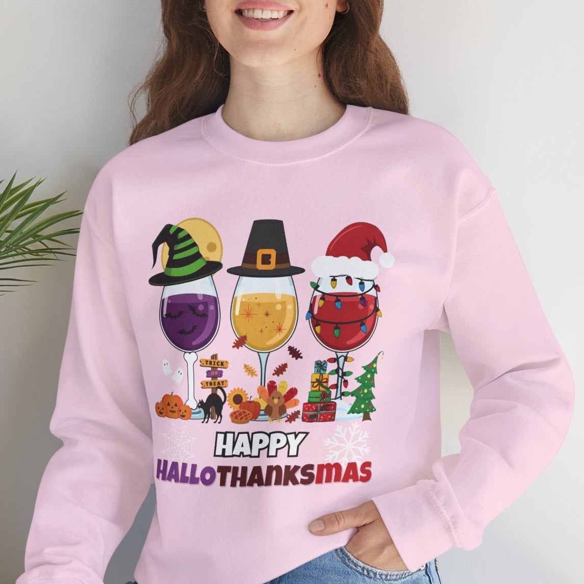 Person wearing a pink sweatshirt with 'Happy Hall-O-THANKSMAZ' design, sitting on a white surface with a plant in the background.