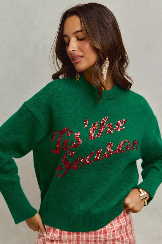 Woman wearing a green sweater with 'Tis the Season' red sequins text on a gray background