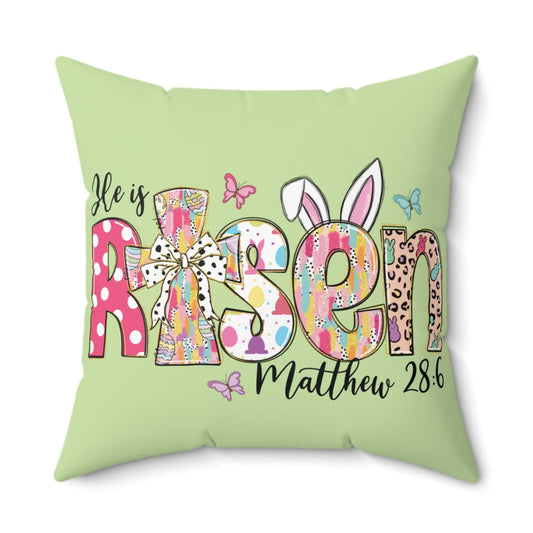 A square throw pillow with a green background featuring a double-sided print with the text 'HE IS RISEN' and decorative elements such as eggs and bunny ears, indicating a spring or Easter theme.
