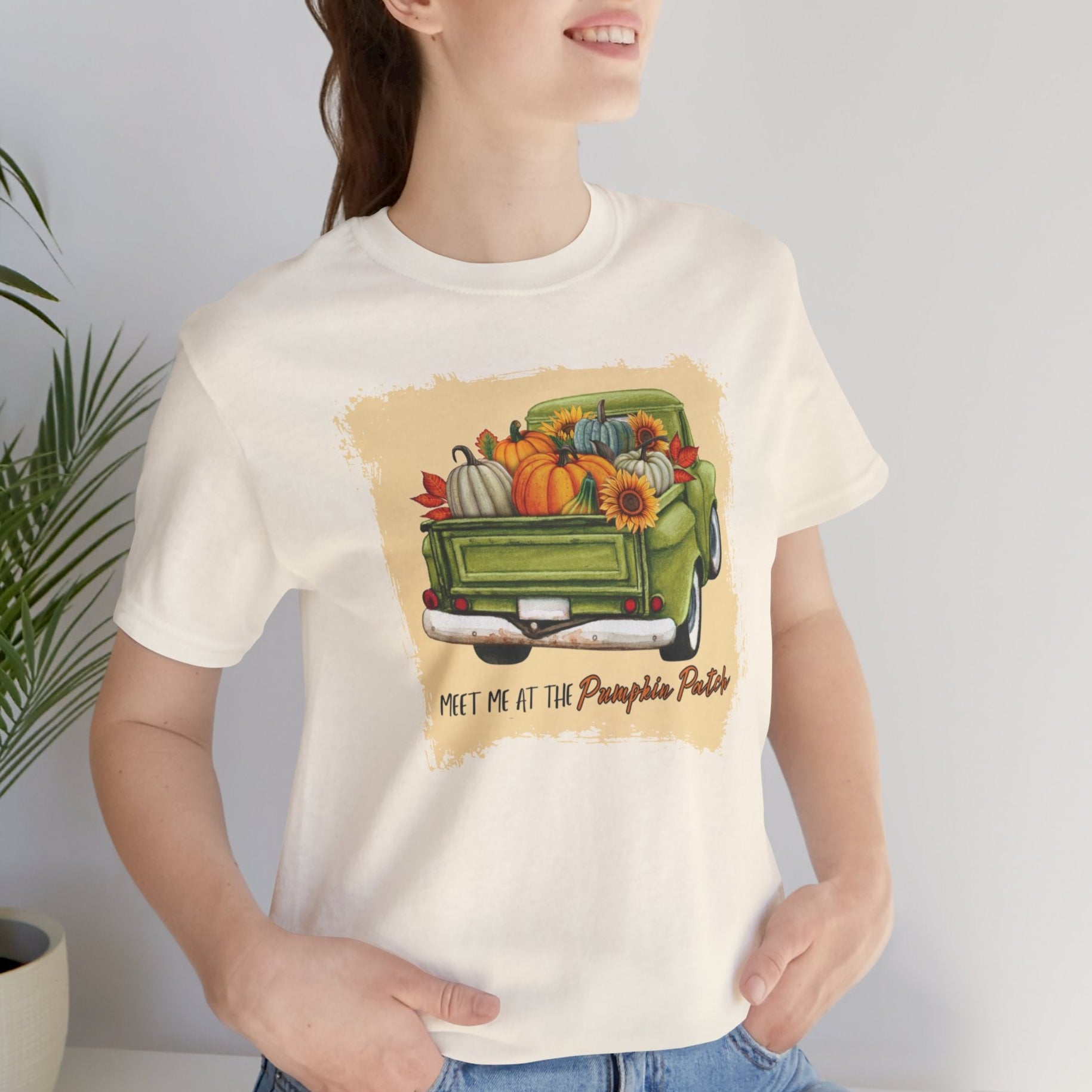 Person wearing a cream t-shirt with a graphic of a green truck carrying pumpkins and fall decorations, with text 'Meet me at the Pumpkin Patch'.