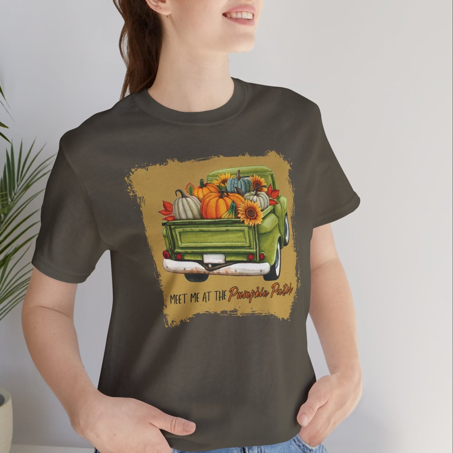 Person wearing a green t-shirt with a graphic of a truck and pumpkins, sitting indoors next to a plant.