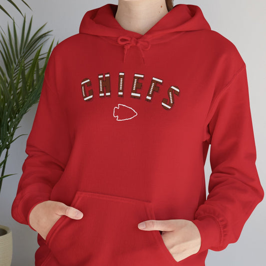 Person wearing a red hoodie with 'CHIEFS' text, with football type font and an arrowhead under the word "CHIEFS" standing indoors next to a plant.
