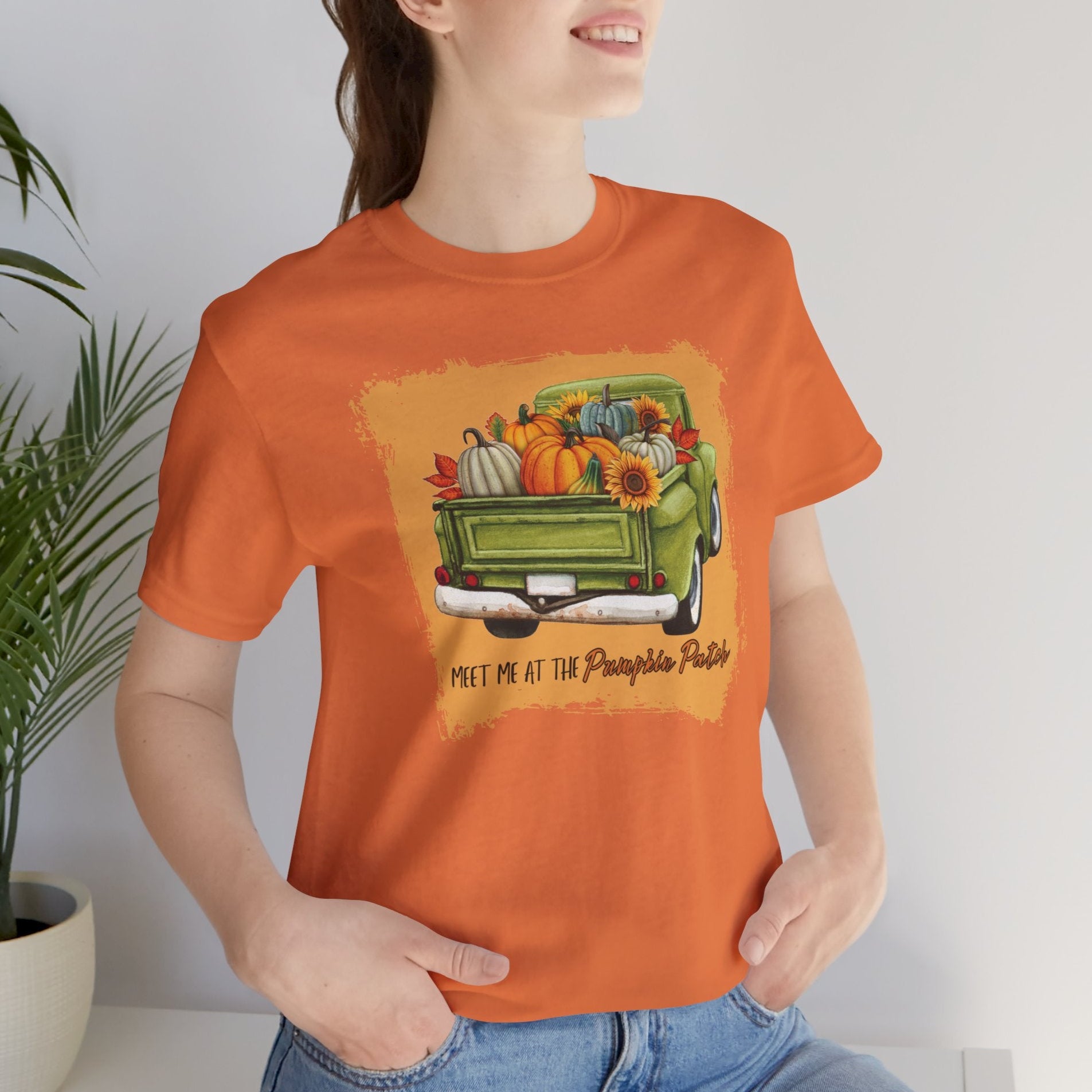 Person wearing an orange t-shirt with a graphic of a truck and pumpkins, sitting indoors.