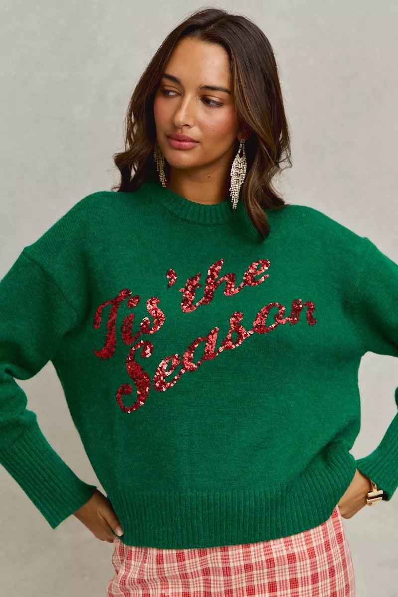 Woman wearing a green sweater with 'Tis the Season' red sequins text on a gray background