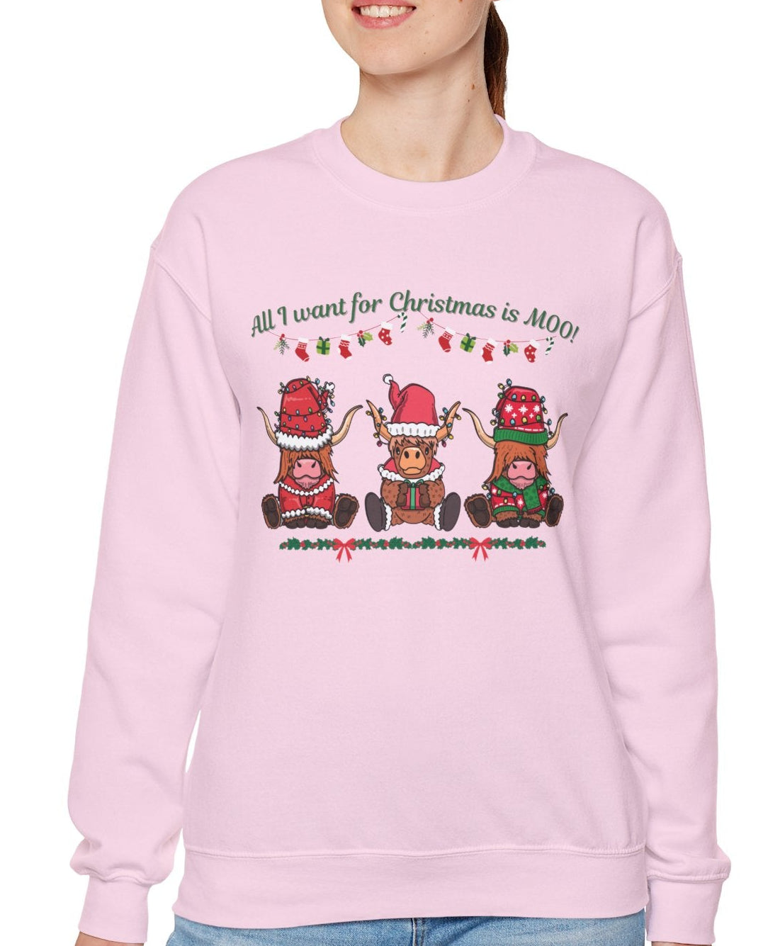 Woman wearing a pink sweatshirt with "All I Want for Christmas Is MOO" sweatshirt featuring three festive cows wearing Santa hats and scarves, printed on cozy crewneck sweaters in white and pink. Exclusive Joyful Verse design by Sara.