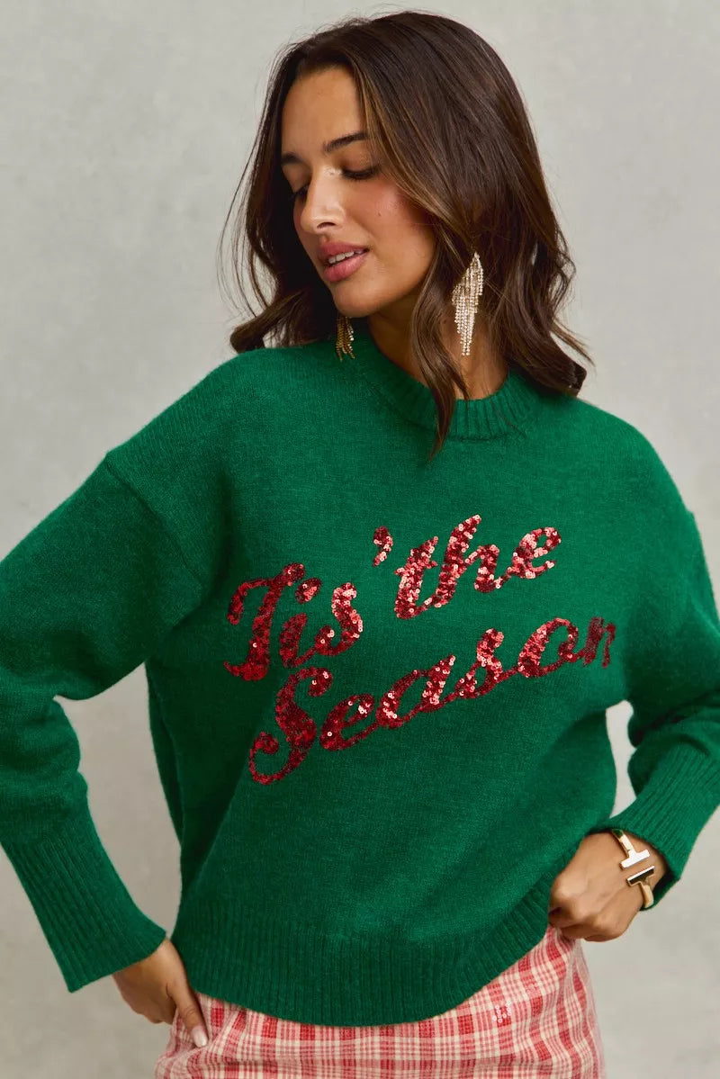Woman wearing a green sweater with 'Tis the Season' red sequins text on a gray background