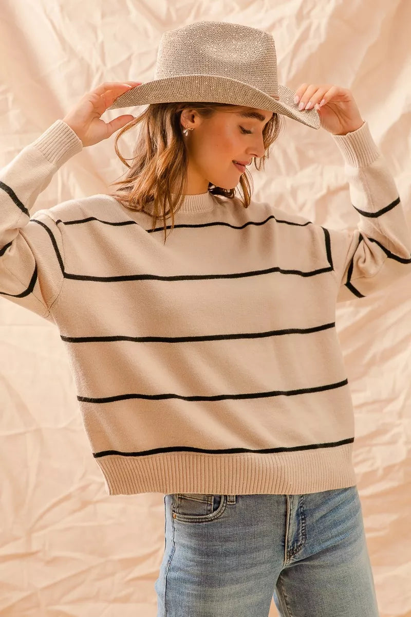 Woman wearing a beige sweater with black stripes and a wide-brimmed hat against a beige background