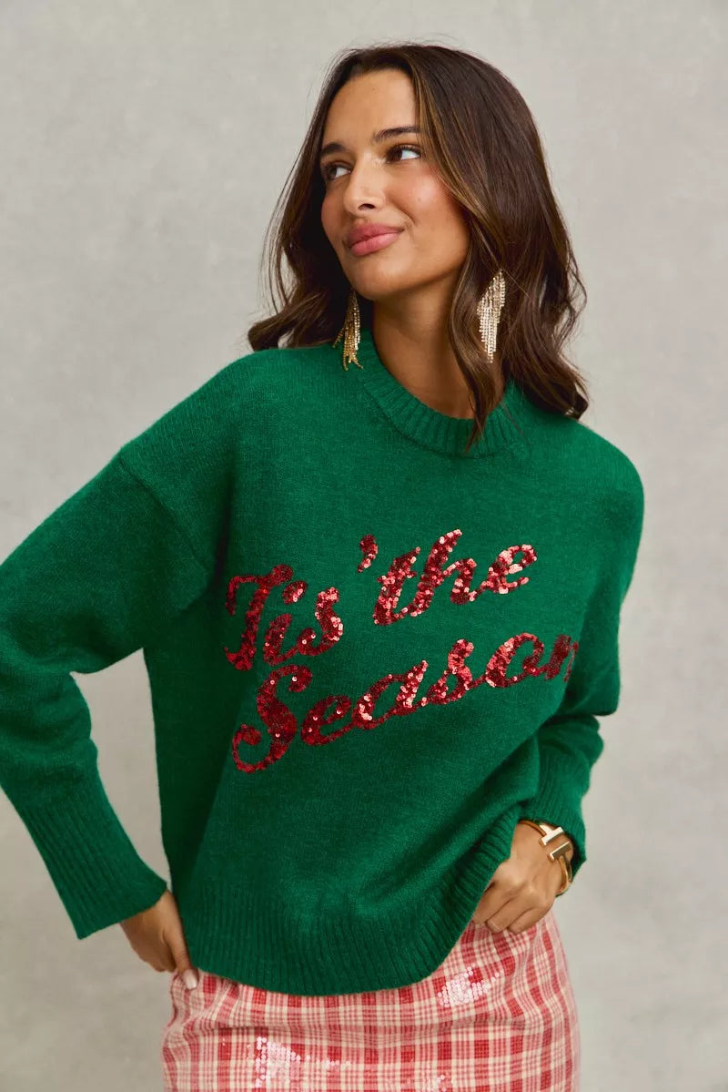 Woman wearing a green sweater with 'Tis the Season' red sequins text on a gray background