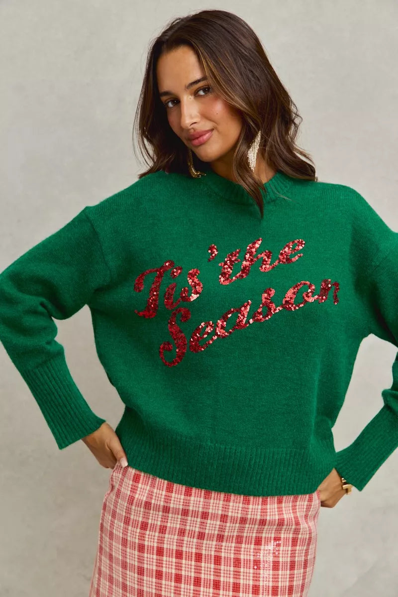 Woman wearing a green sweater with 'Tis the Season' red sequins text on a gray background