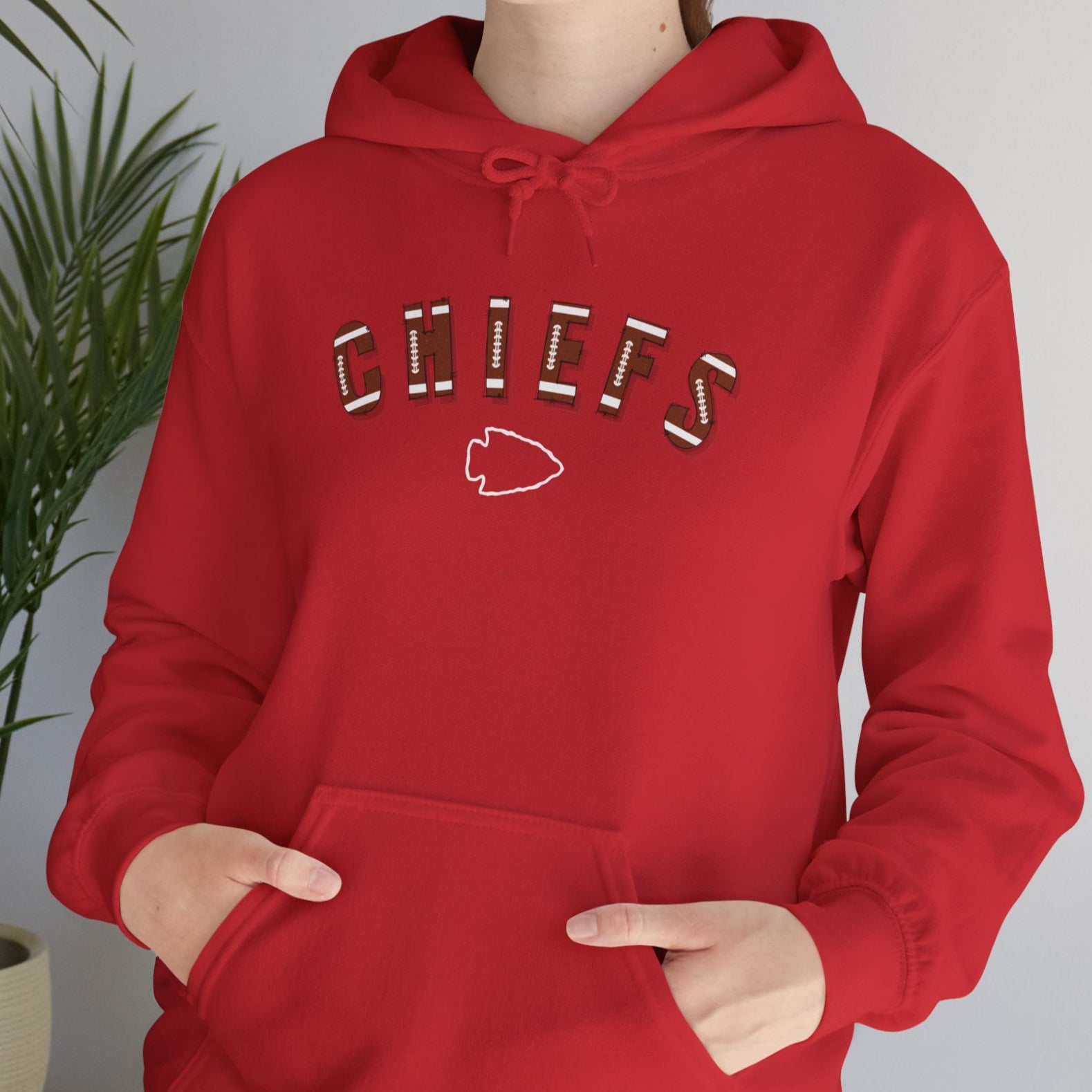 Person wearing a red hoodie with 'CHIEFS' text, with football type font and an arrowhead under the word "CHIEFS" standing indoors next to a plant.