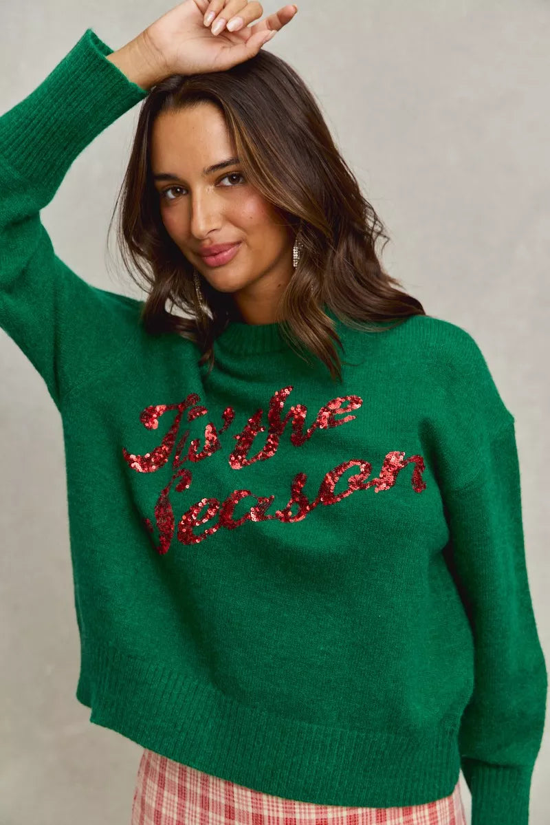 Woman wearing a green sweater with 'Tis the Season' red sequins text on a gray background